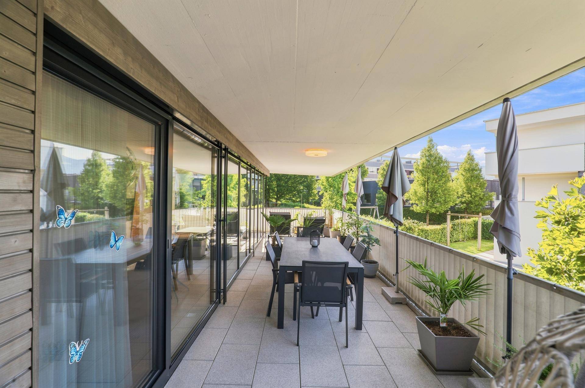 Große, geflieste Terrasse mit Esstisch, Sonnenschirmen und Blick ins Grüne.