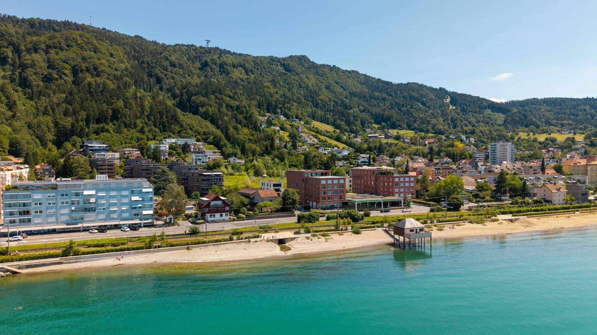 Panoramablick auf die Küstenstadt mit Seeufer, Strand und bewaldeten Bergen im Hintergrund an einem sonnigen Tag.