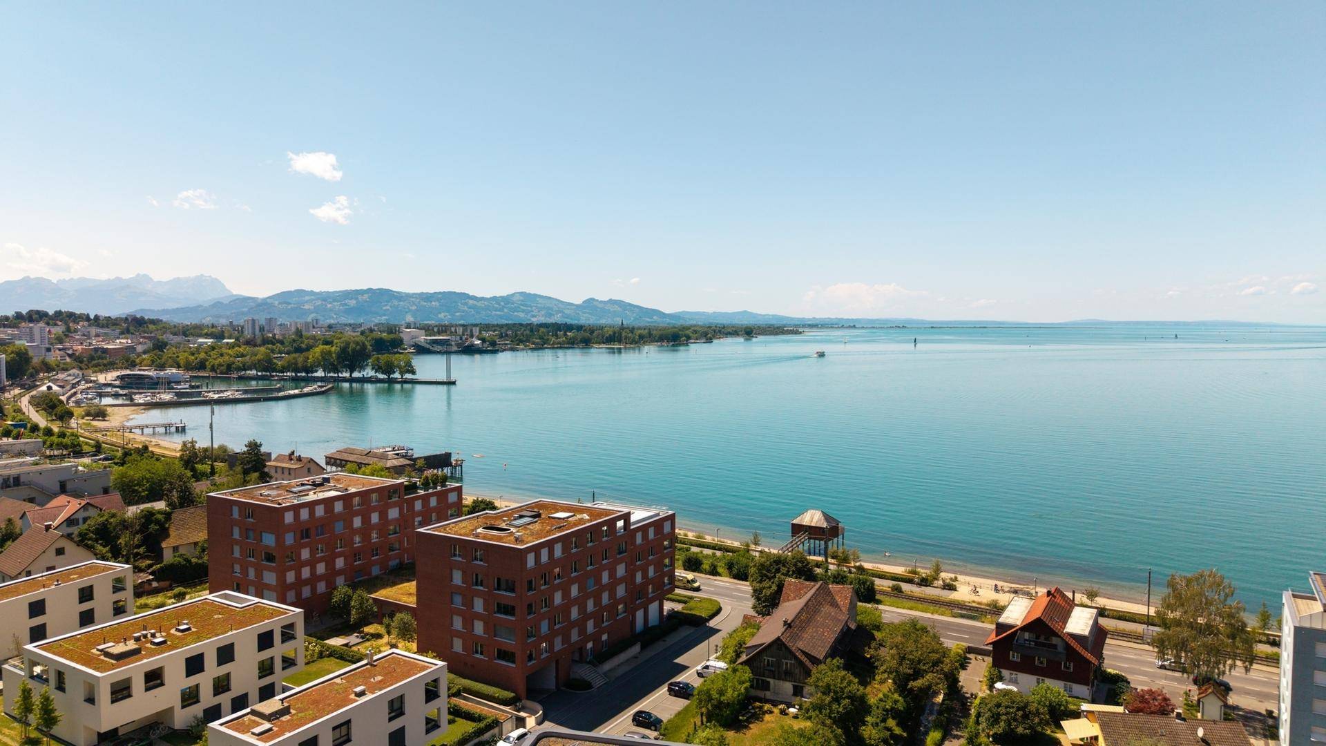 Weitläufiger Blick über den See und die Küstenlinie mit modernen Wohngebäuden und Bergen im Hintergrund.