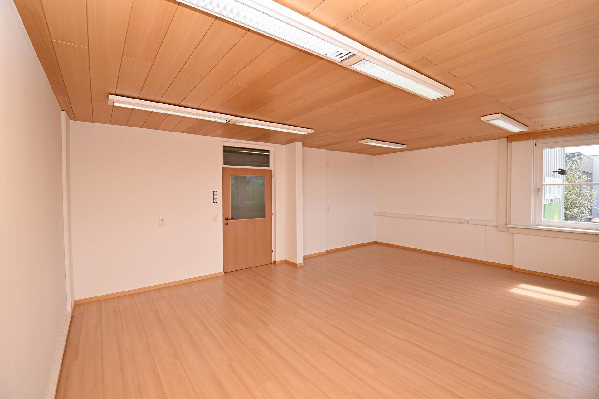 Heller Büroraum mit Laminatboden, Holzdecke und Zugang zu weiteren Bereichen.
