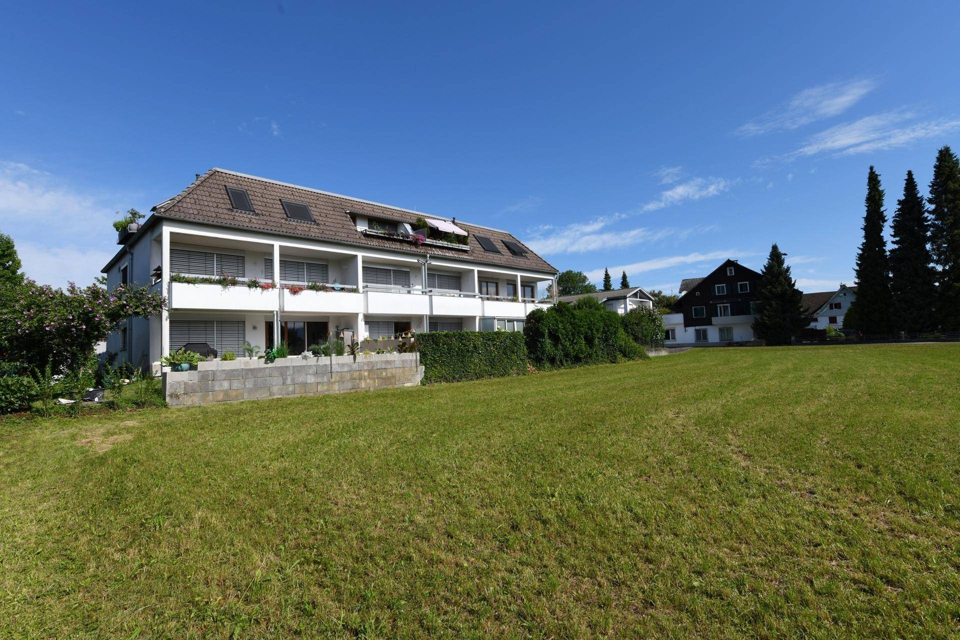 Mehrfamilienhaus mit mehreren Balkonen und großer Grünfläche unter blauem Himmel.