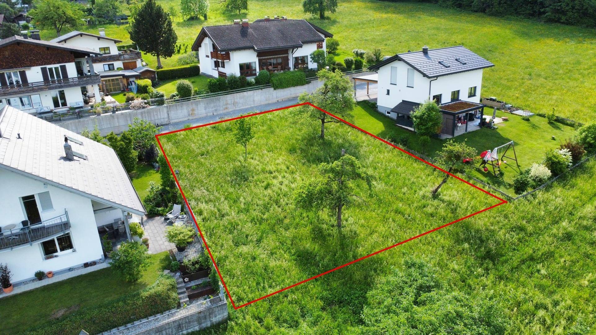 Luftaufnahme eines grünen Baugrundstücks mit roter Umrandung, umgeben von Wohnhäusern und Natur.