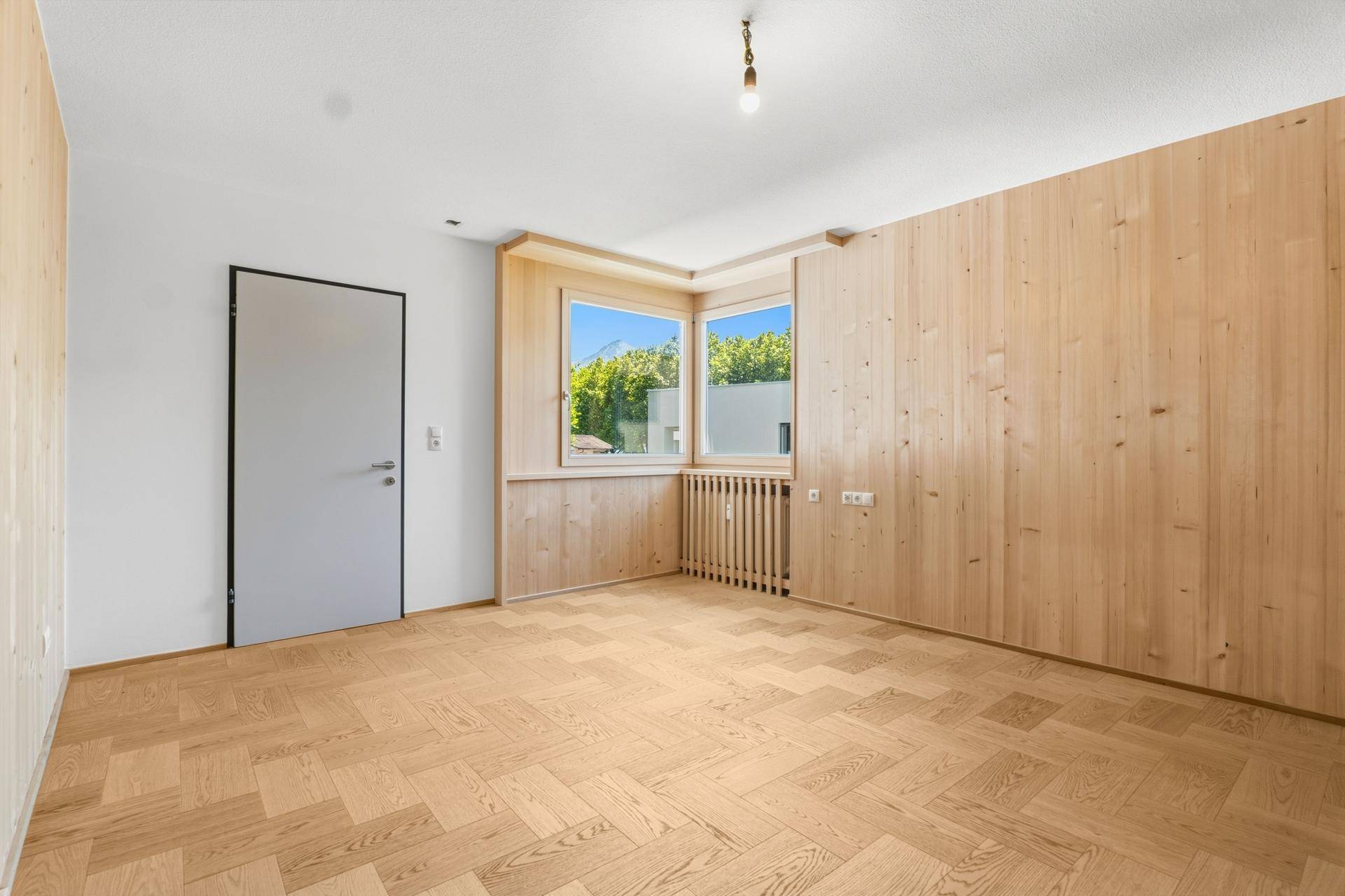 Heller Raum mit Holzwandverkleidung, Fischgrätparkett und zwei Fenstern.