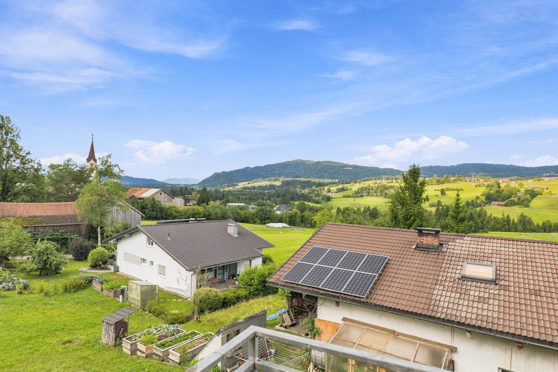 Weitläufiger Blick über die grüne Landschaft mit Häusern, Feldern und einem Kirchturm in der Ferne.
