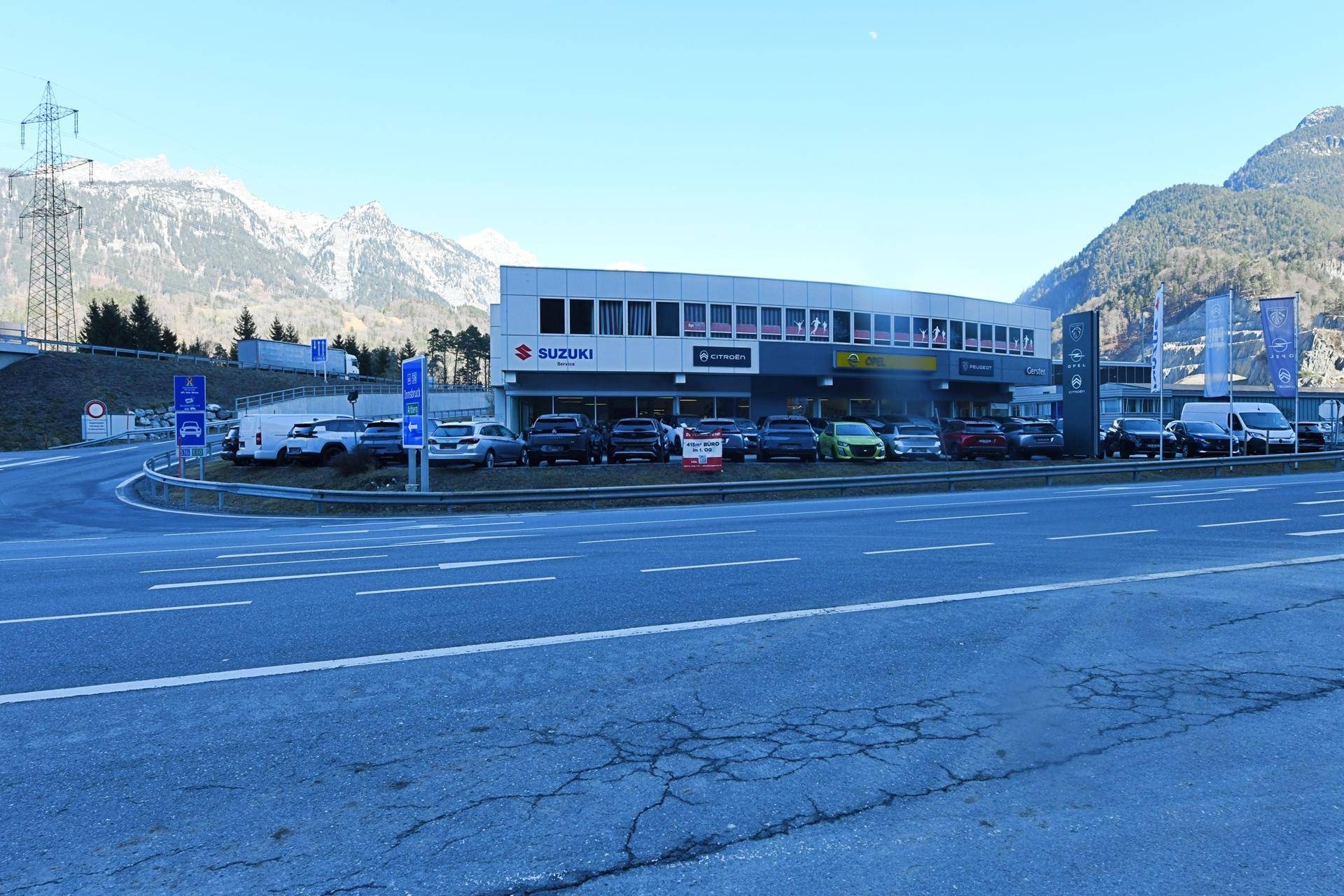 Außenansicht eines Autohauses mit Parkplätzen und Bergen im Hintergrund unter blauem Himmel.