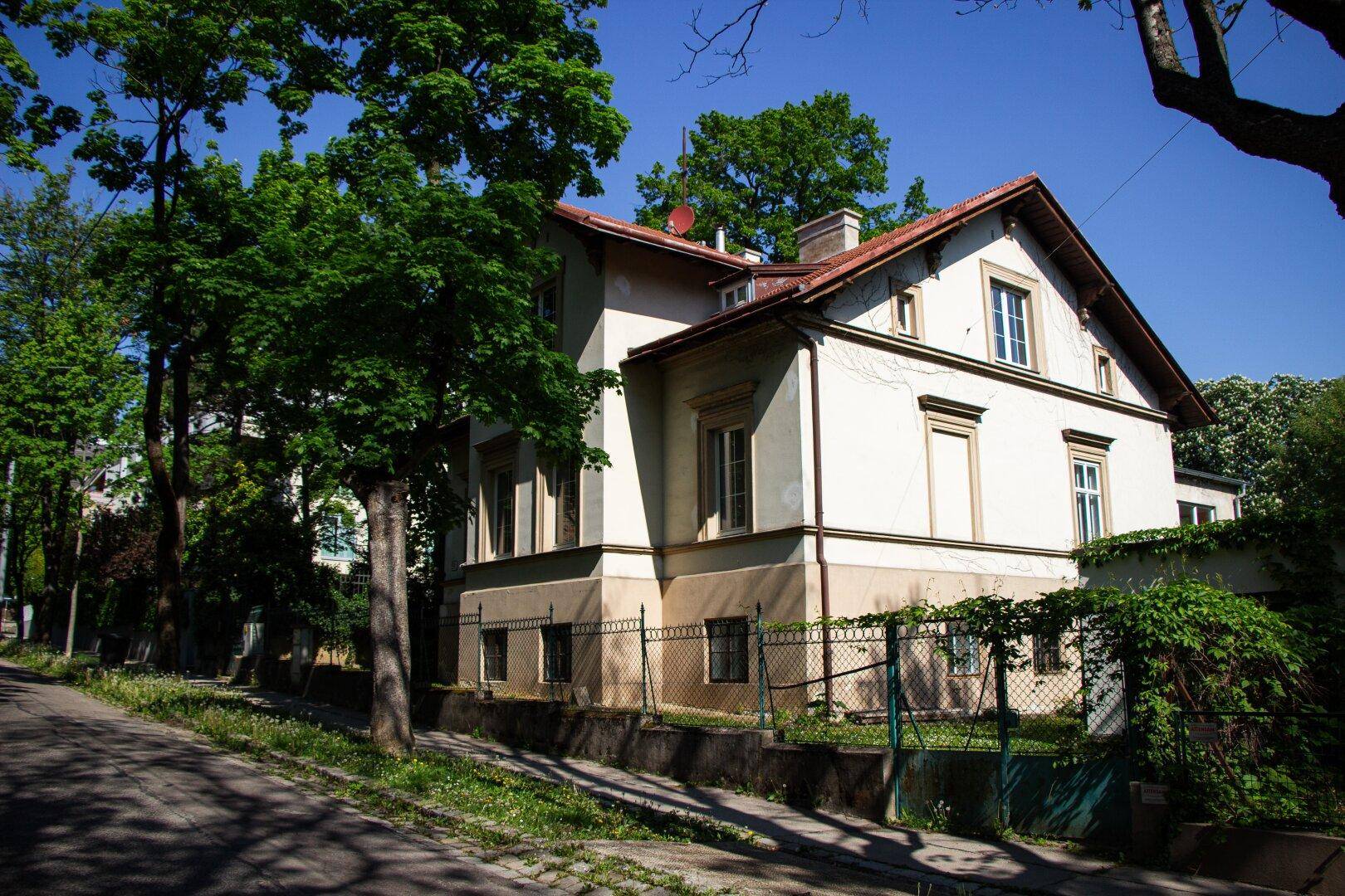 Eckansicht des Wohnhauses von der Straße, umgeben von Bäumen und einem Zaun.