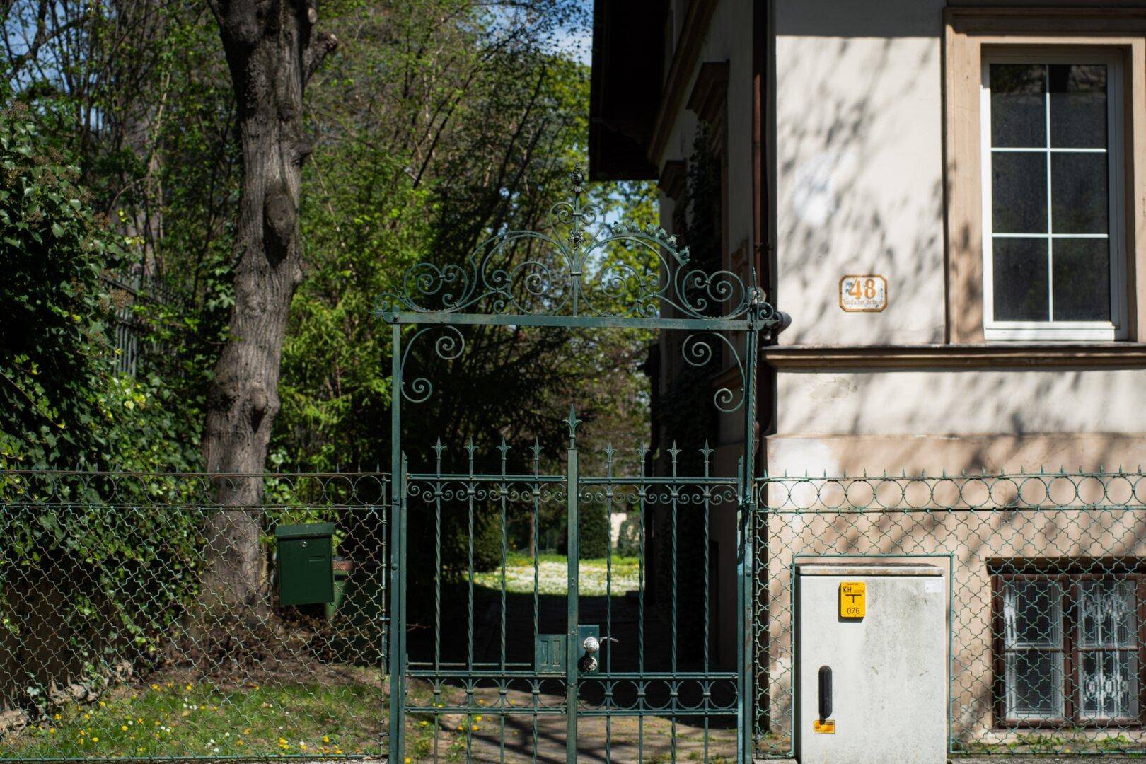 Schmiedeeisernes Eingangstor zum Grundstück mit einem Teil der Hausfassade und üppigem Grün.