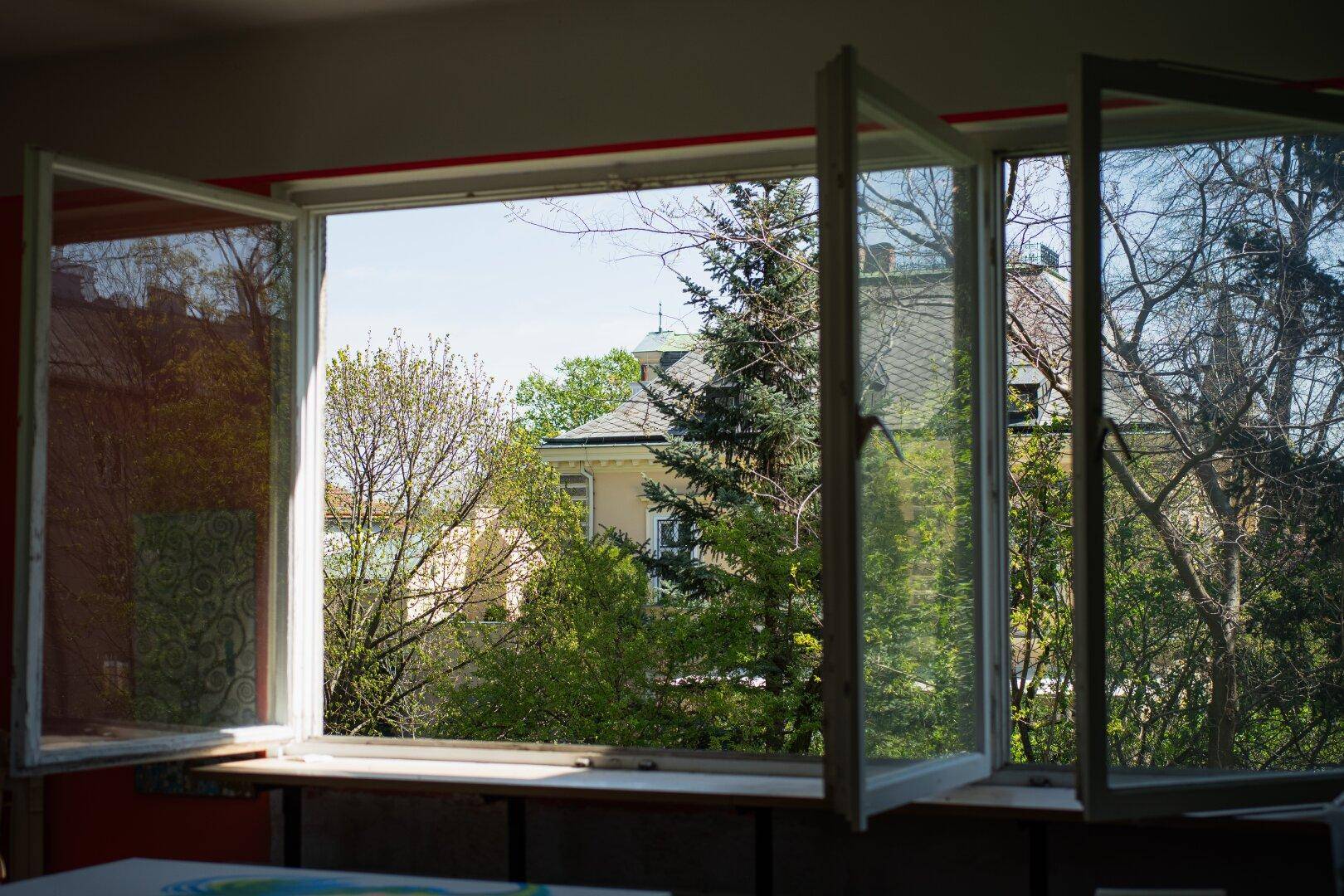 Blick aus einem geöffneten Fenster auf grüne Bäume und umliegende Häuser.