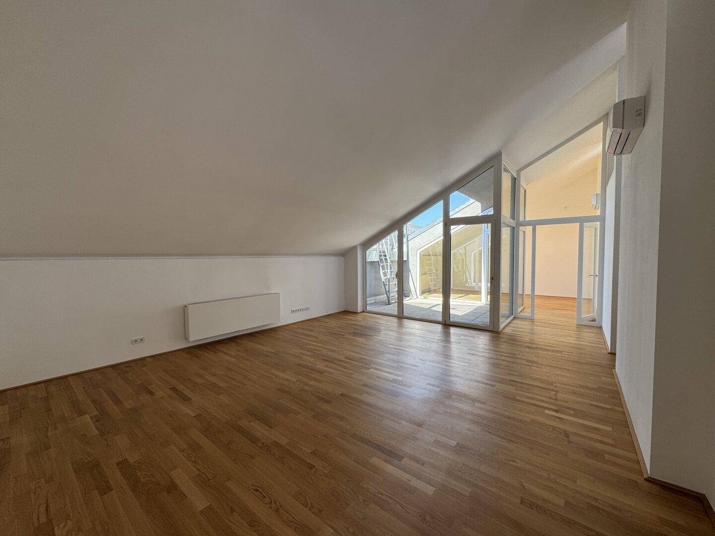 Geräumiges, leeres Wohnzimmer mit hellem Parkettboden und viel Tageslicht durch die Glasfronten.