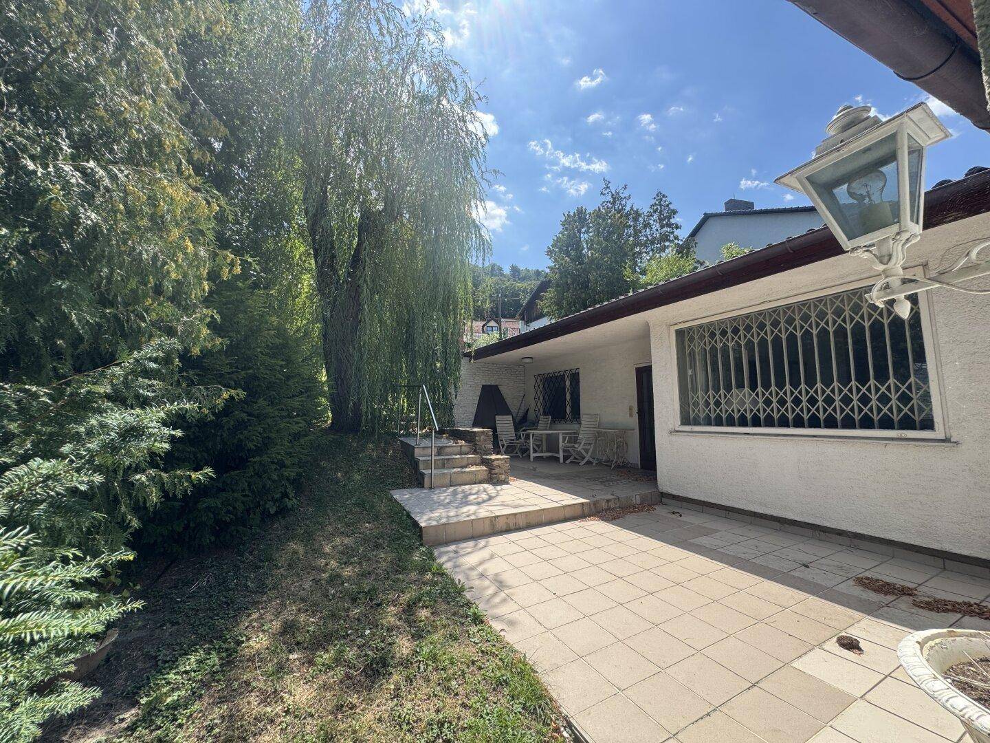 Gepflasterte Terrasse mit Gartenmöbeln und einem großen Baum, ideal für Entspannung im Freien.