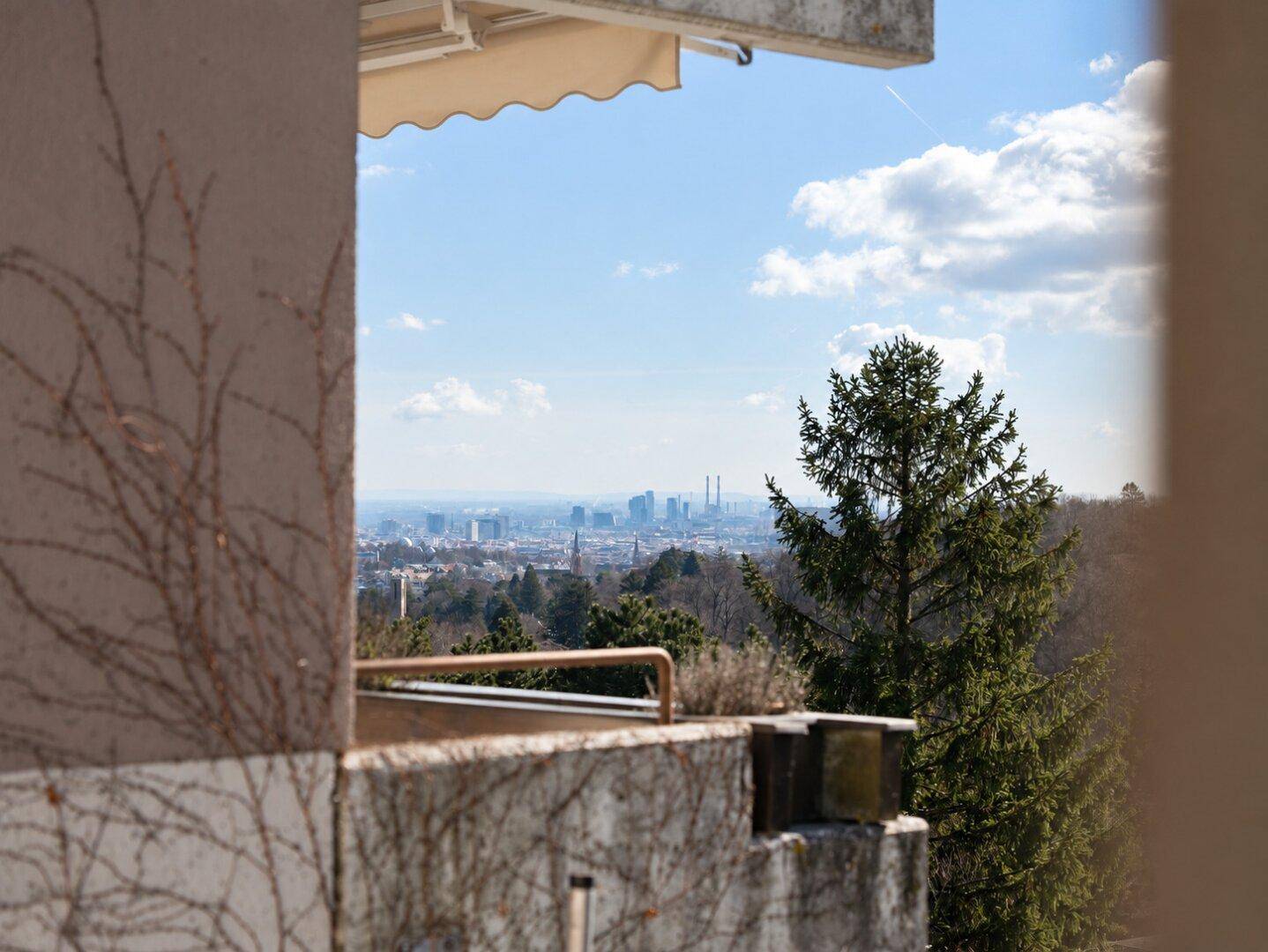 Weitblick von einem Balkon auf eine Stadtlandschaft und grüne Hügel unter blauem Himmel.