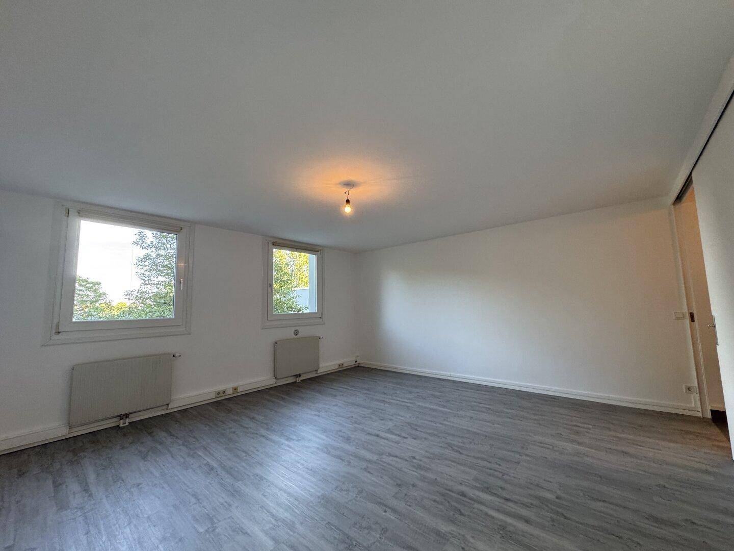 Geräumiger, heller Raum mit grauem Laminatboden und zwei Fenstern, ideal als Wohn- oder Schlafzimmer.