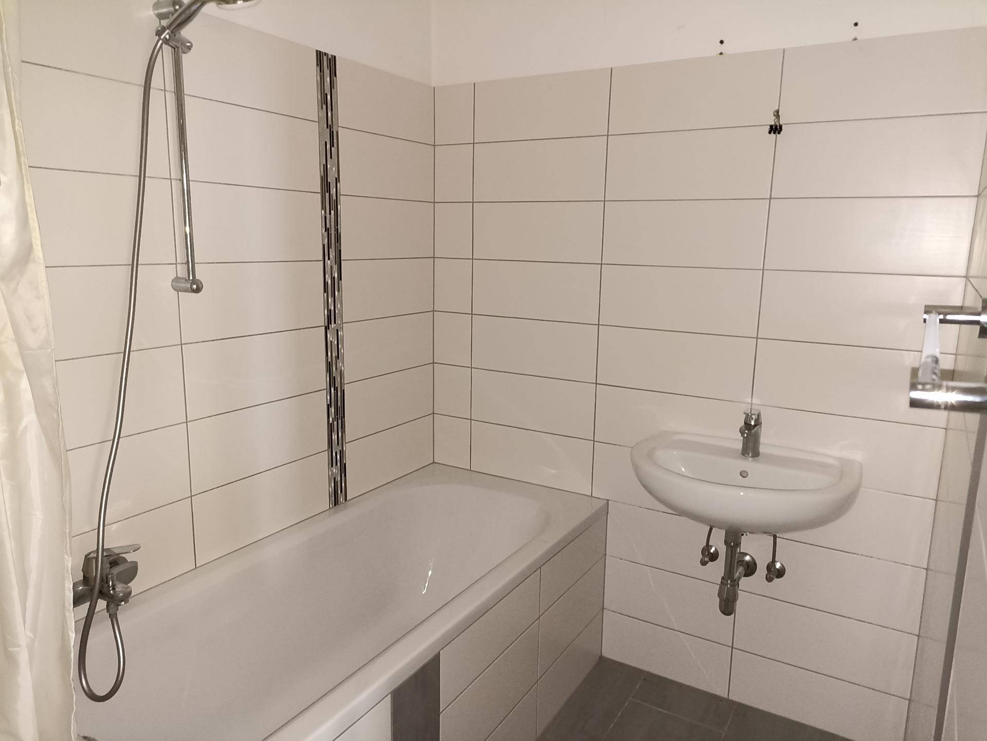 Helles Badezimmer mit Badewanne, Handbrause und einem kleinen Waschbecken an der Wand.