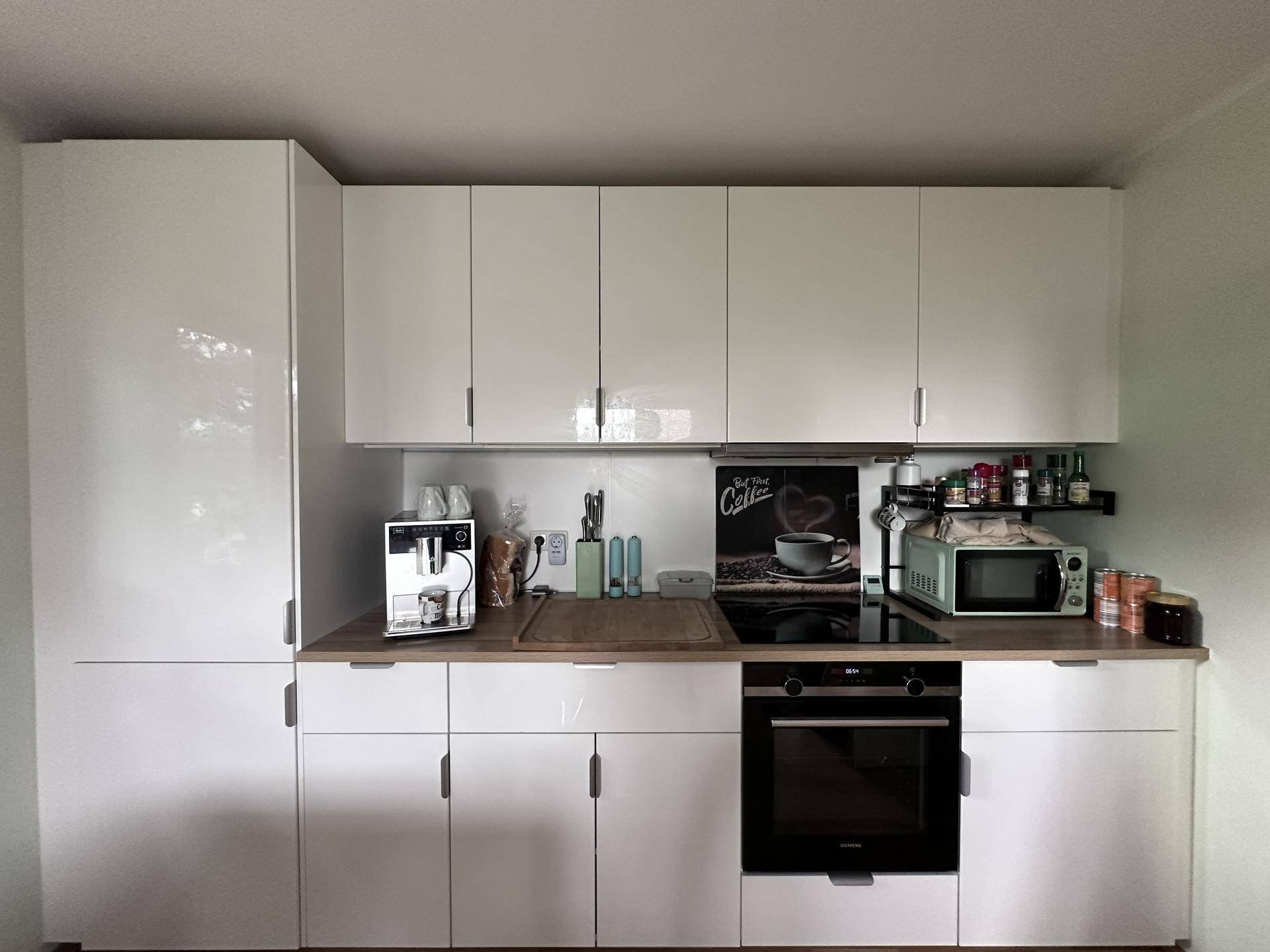Kompakte, moderne Küche mit weißen Hochglanzfronten, integriertem Backofen und Kaffeemaschine.