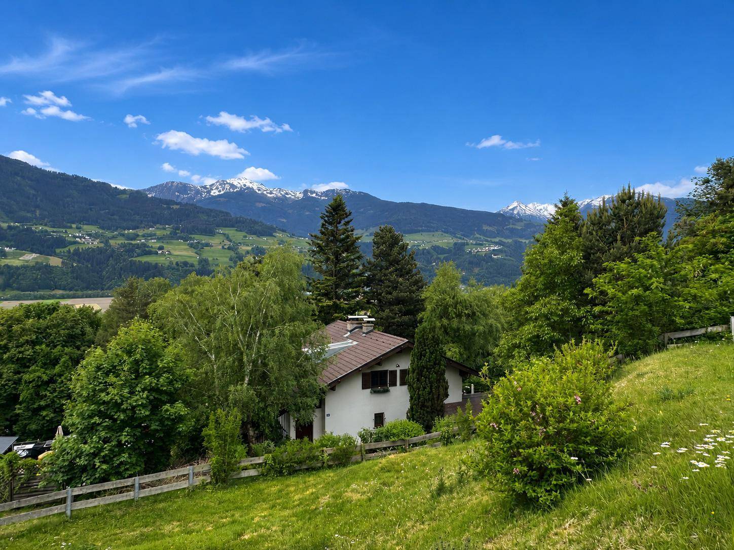 Charmantes Haus in ländlicher Umgebung mit grünen Wiesen und schneebedeckten Bergen im Hintergrund.