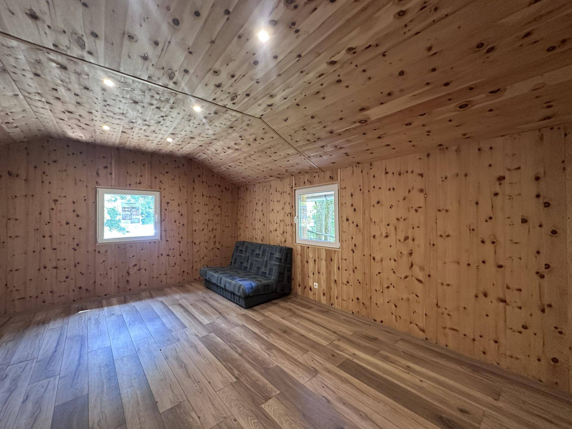 Gemütliches Wohnzimmer mit Holzvertäfelung, Schlafsofa und zwei Fenstern für Tageslicht.