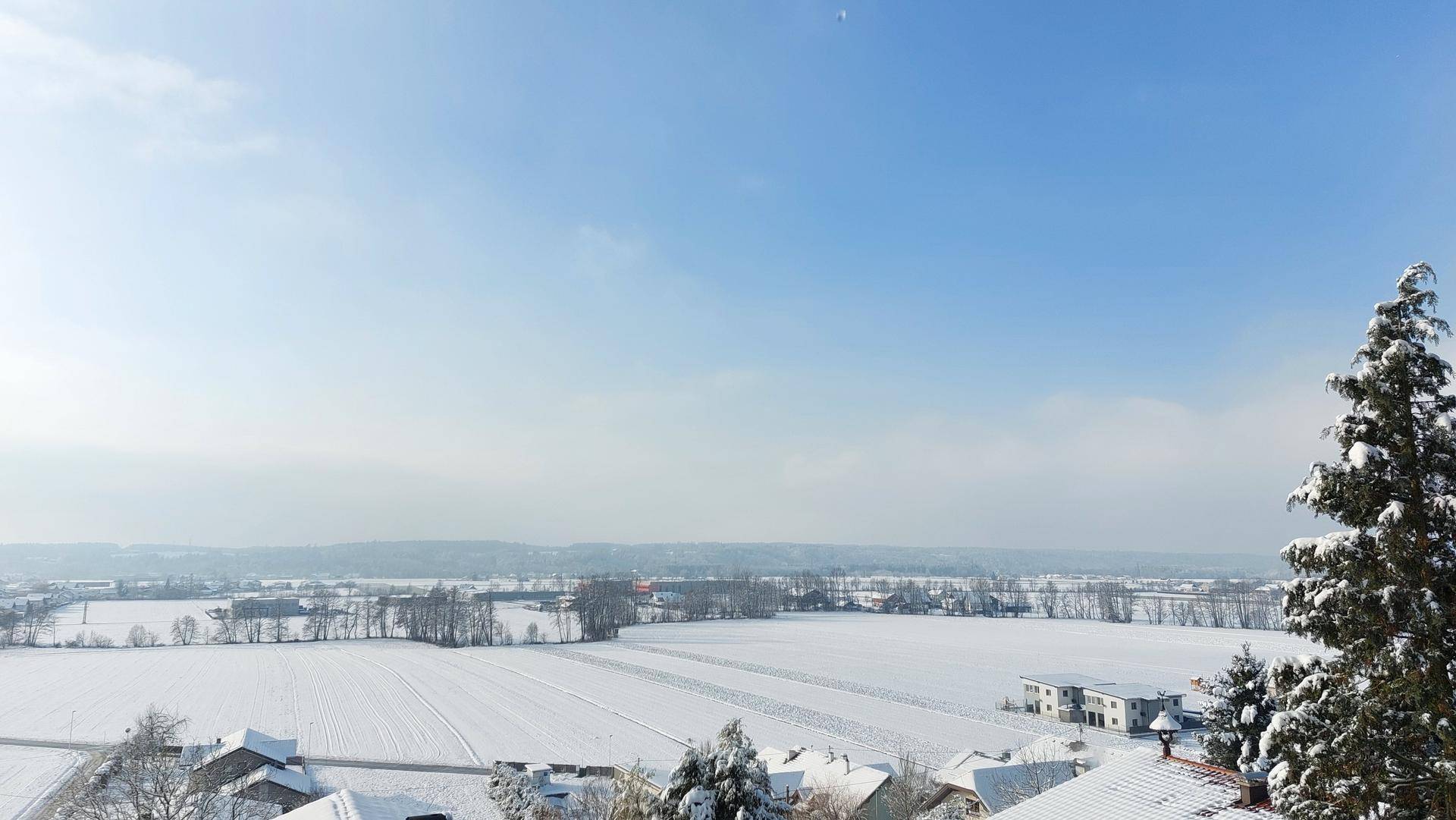Winterliche Landschaft mit schneebedeckten Feldern, Häusern und einem weiten Blick auf die Ferne unter blauem Himmel.