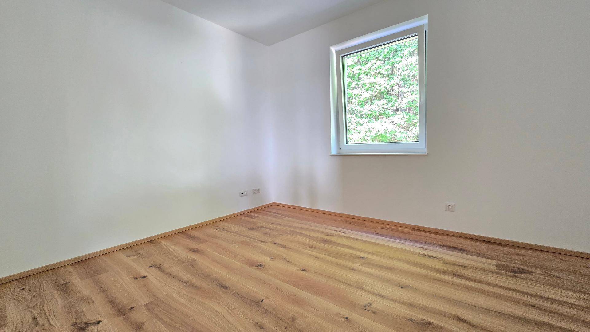 Helles Schlafzimmer mit Holzboden und einem Fenster, das einen angenehmen Blick ins Grüne bietet.