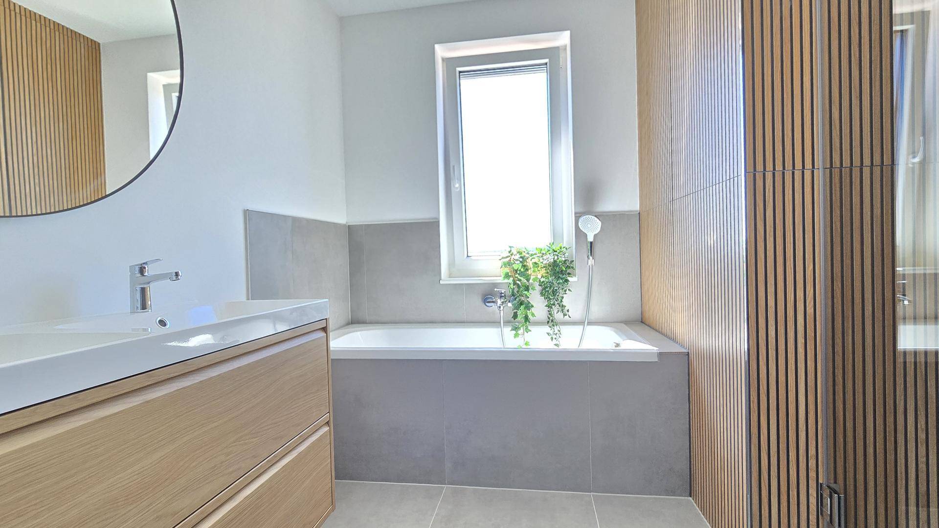 Modernes Badezimmer mit Badewanne, Holzlamellenwand und Fenster für natürliches Licht.