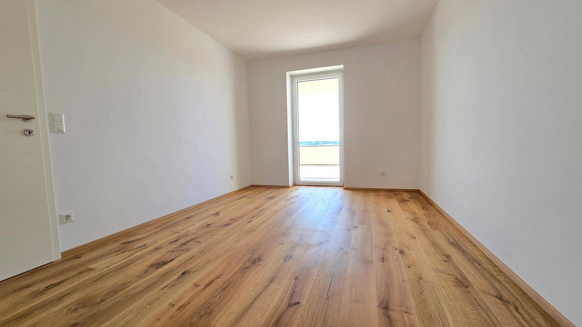 Geräumiges, leeres Zimmer mit Holzplankenboden und einer Fenstertür, die viel Tageslicht hereinlässt.