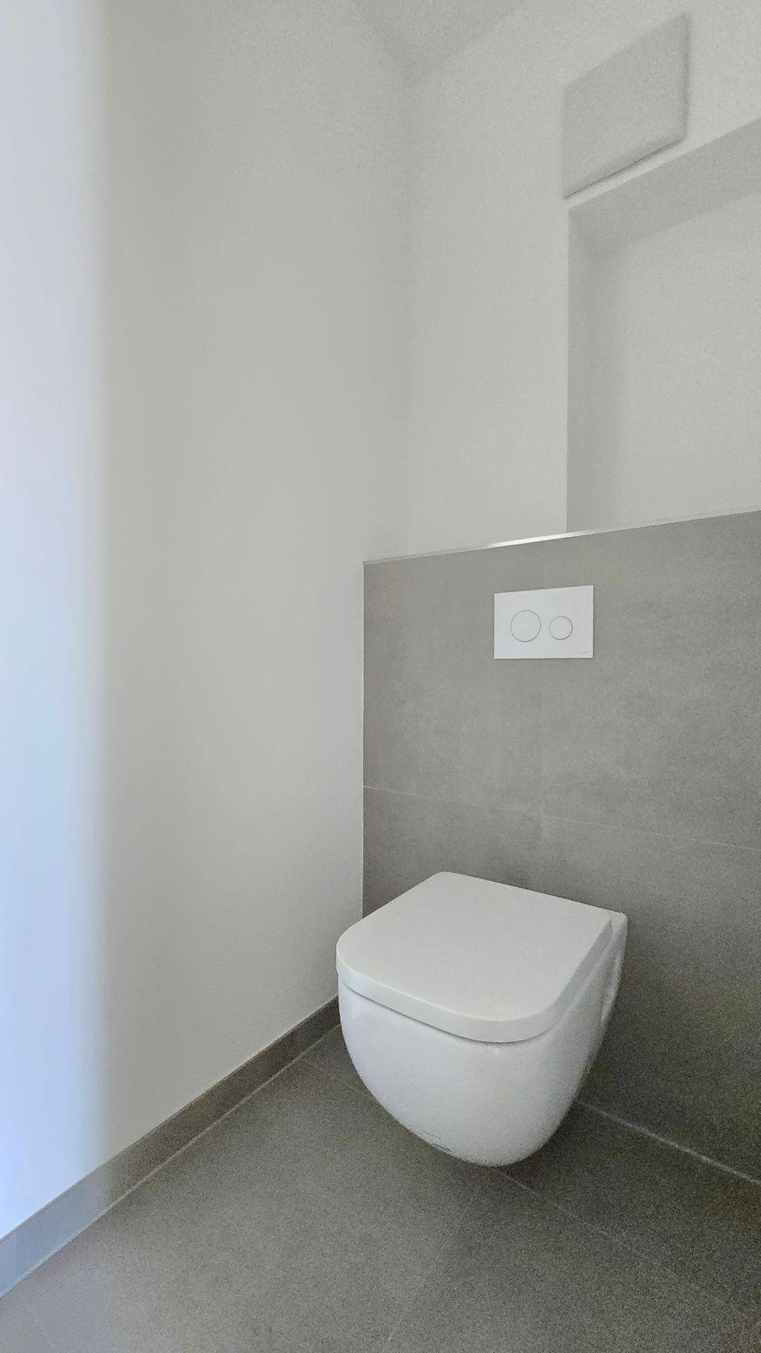 Modernes WC mit wandmontierter Toilette und grauen Fliesen an der Wand, minimalistisches Design.