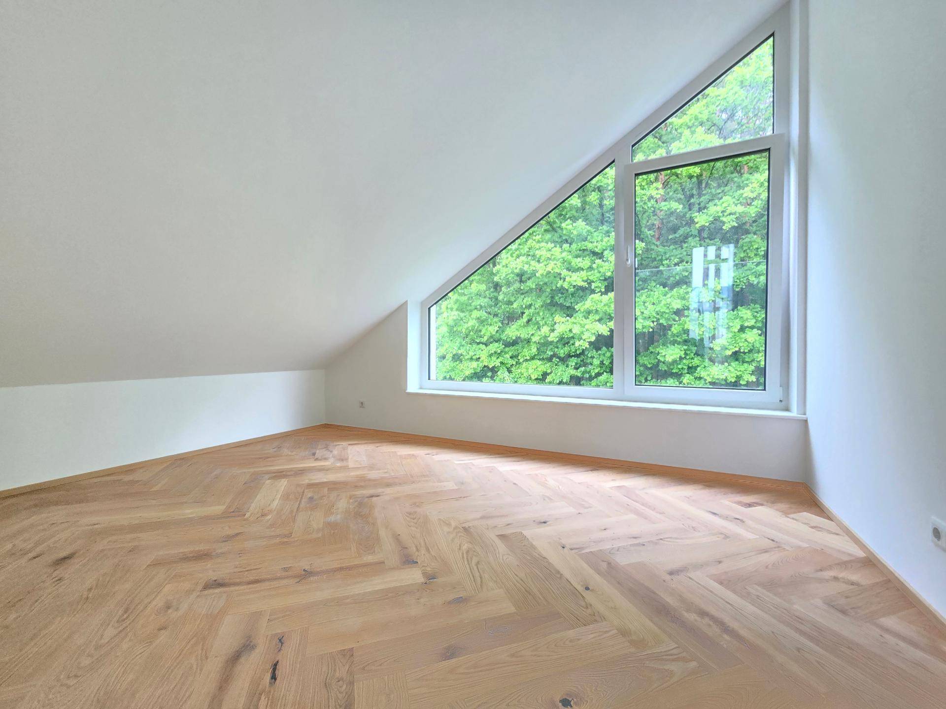 Helles Zimmer im Dachgeschoss mit Fischgrätparkett und großem dreieckigem Fenster mit Grünblick.