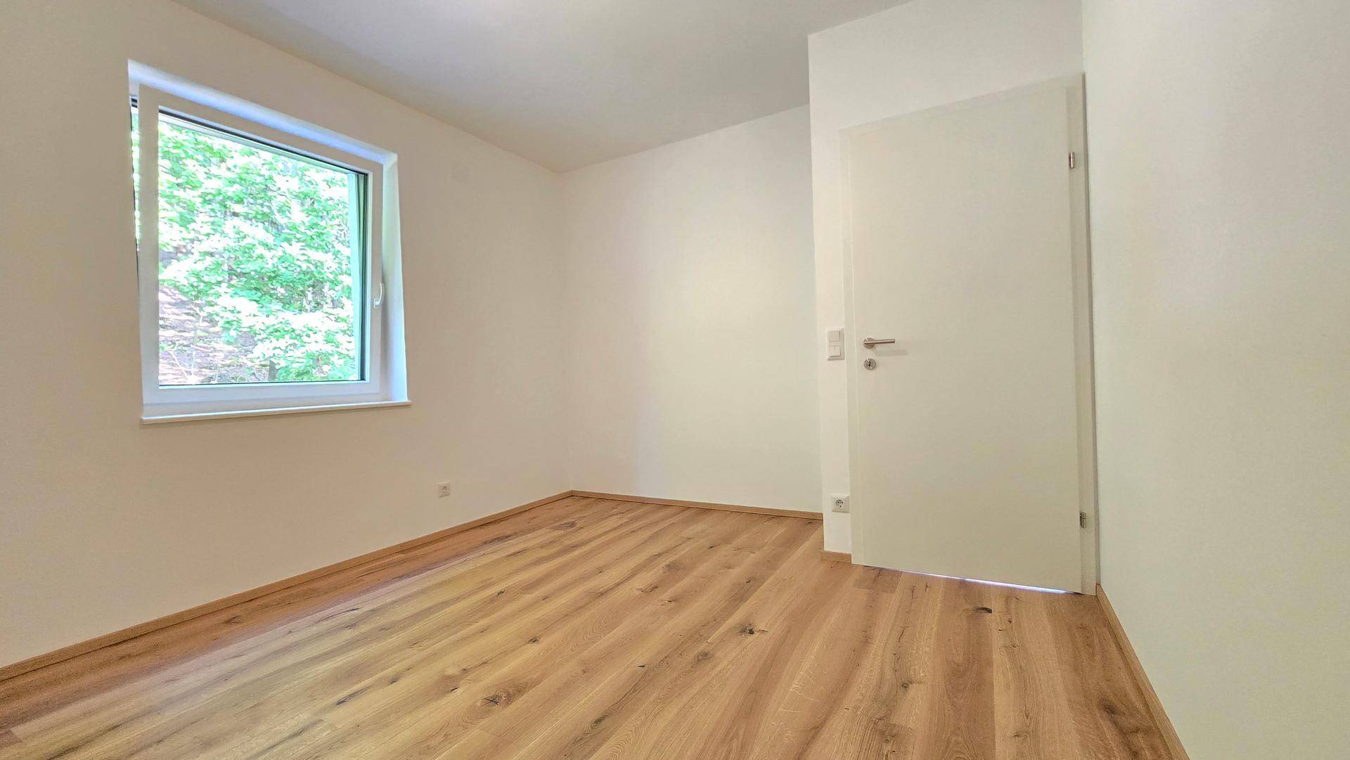 Leeres Schlafzimmer mit Holzboden und weißer Tür, gut beleuchtet durch das Fenster mit Grünblick.