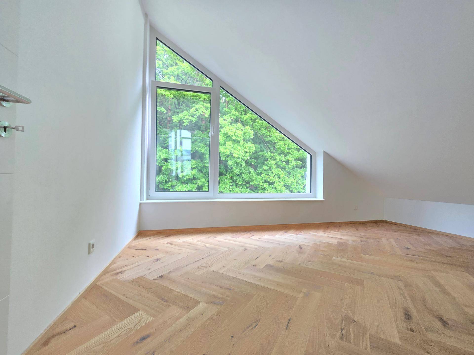 Dachgeschosszimmer mit Fischgrätparkett und großem Fenster, das einen schönen Blick ins Grüne bietet.