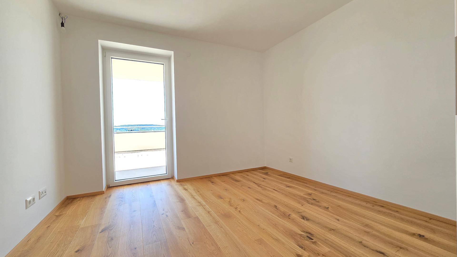 Leeres Schlafzimmer mit hellem Holzplankenboden und Zugang zu einem Außenbereich durch eine Fenstertür.