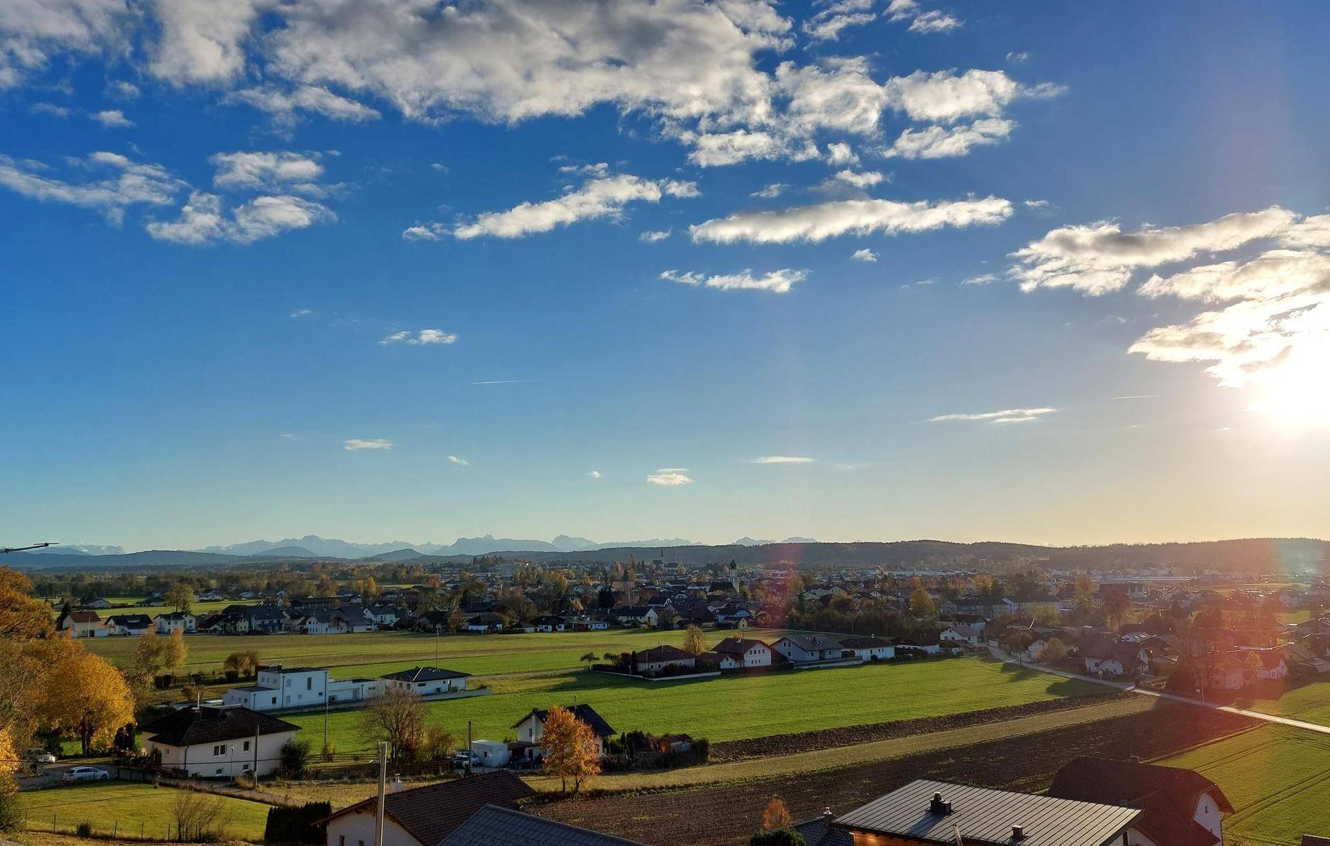 Weitläufiger Blick über die ländliche Umgebung mit Häusern, Feldern und fernen Bergen unter blauem Himmel.