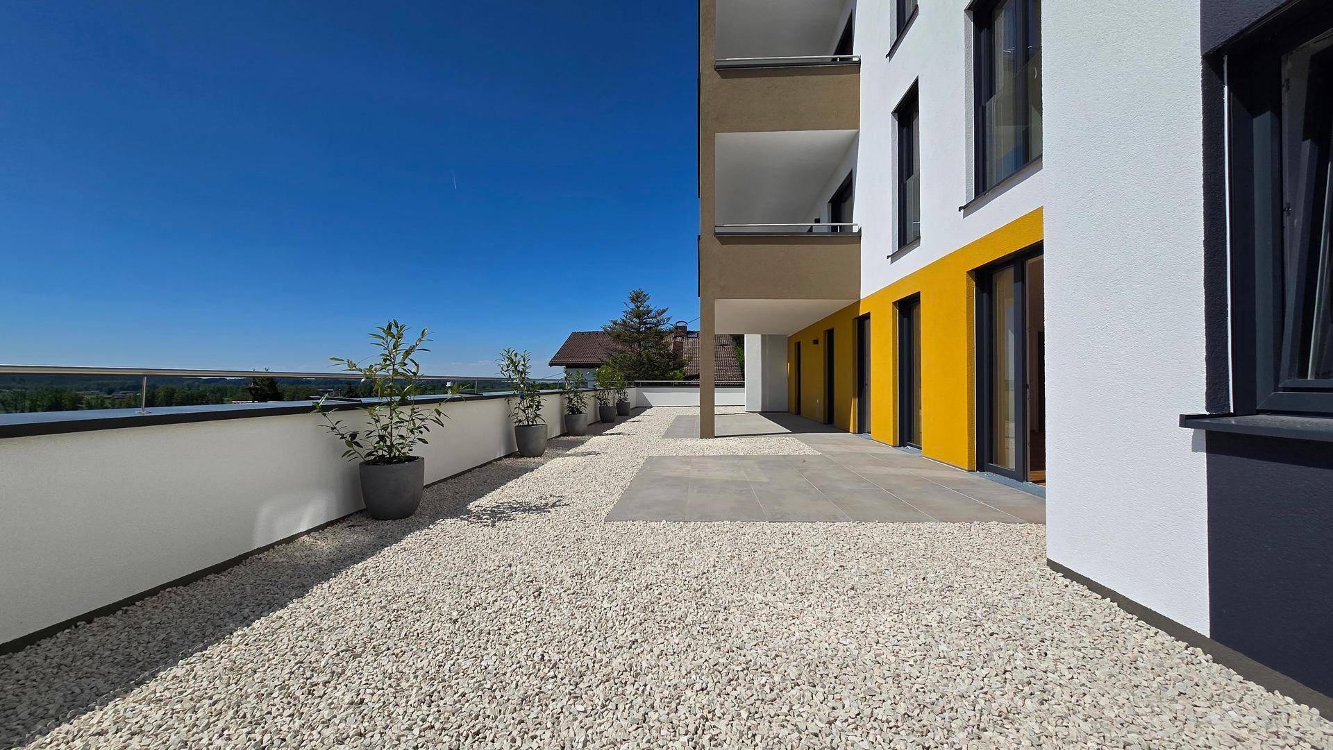 Weitläufige Terrasse mit Kies und Fliesen, angrenzend an die moderne Gebäudefassade mit gelben Akzenten.