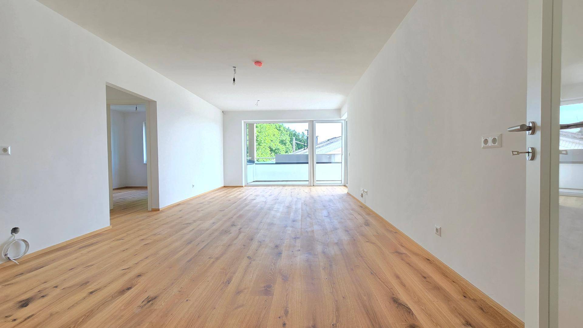 Helles, leeres Wohnzimmer mit neuem Holzboden und großem Fenster mit Zugang zu einem Balkon.