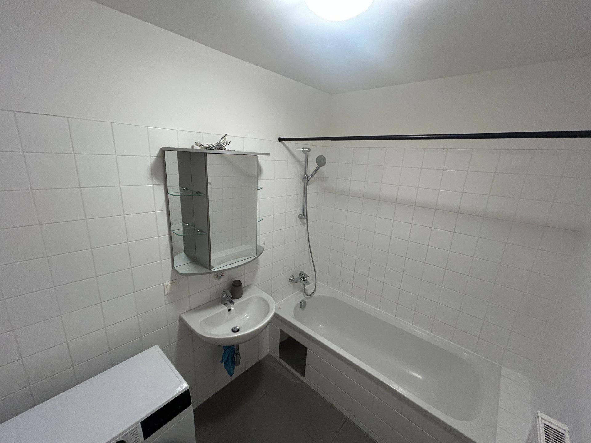 Helles Badezimmer mit weißen Fliesen, Badewanne mit Duschfunktion und Waschbecken.