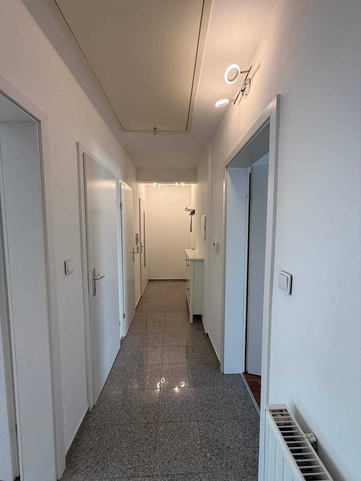 Langer Flur mit Fliesenboden, weißen Türen und Deckenbeleuchtung.