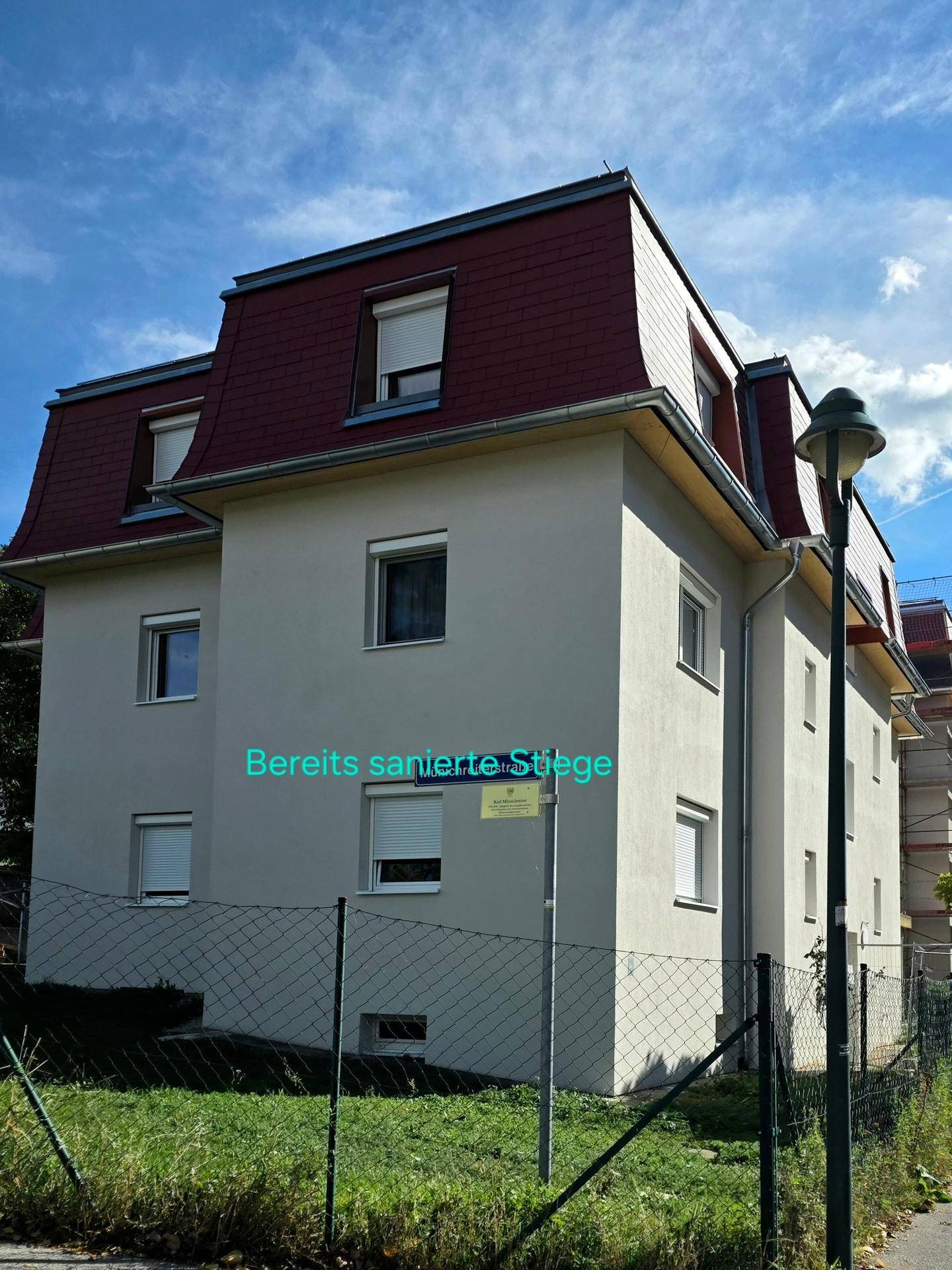Renoviertes Mehrfamilienhaus mit Mansarddach und gepflegter Fassade unter blauem Himmel, von der Seite gesehen.