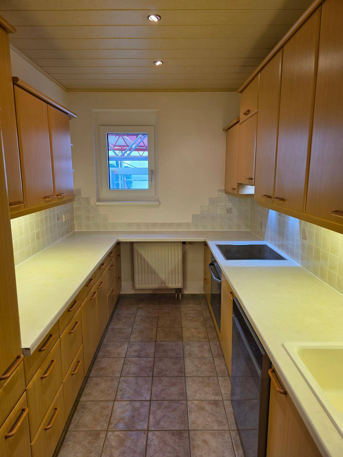 Funktionale Küche mit Holzschränken, hellen Arbeitsplatten und Fliesenboden, gut beleuchtet durch ein Fenster.