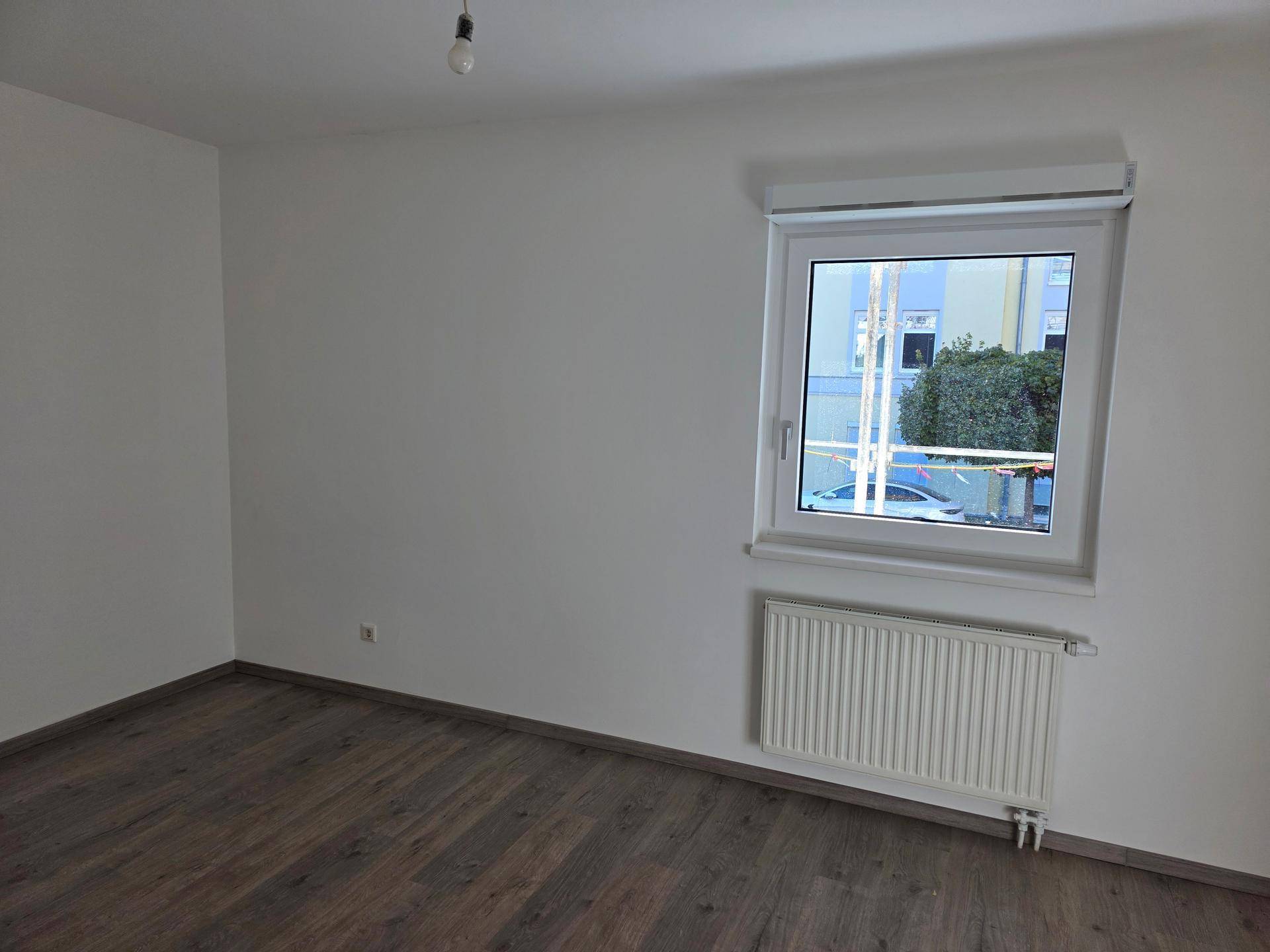 Helles Zimmer mit dunklem Holzboden und Fenster, ausgestattet mit einem Rollladen für Privatsphäre und Komfort.
