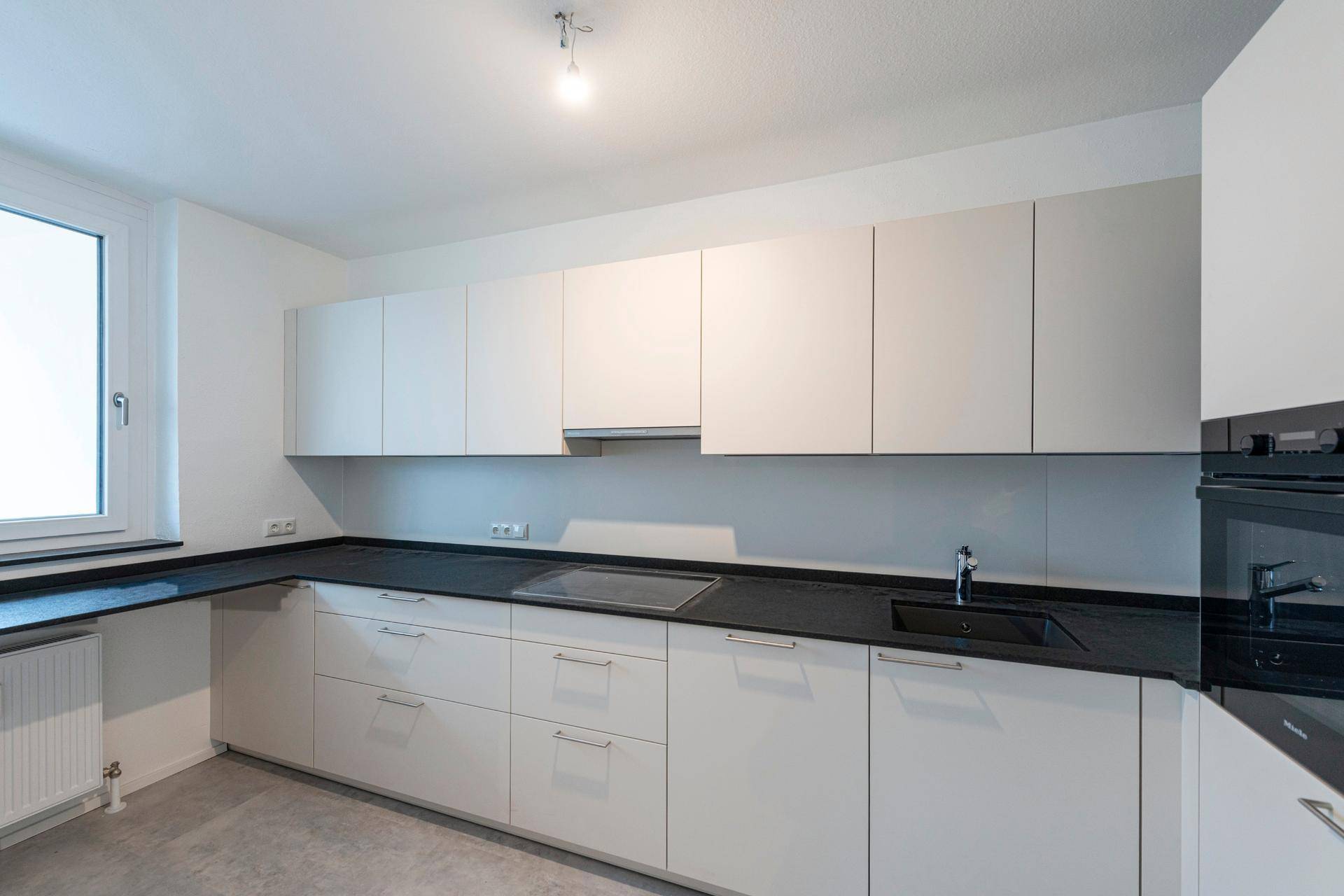 Moderne Einbauküche mit weißen Schränken, dunkler Arbeitsplatte und Fenster.