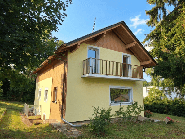 Freistehendes Einfamilienhaus mit gelber Fassade, Balkon und umgebendem Grün, bei Sonnenschein.