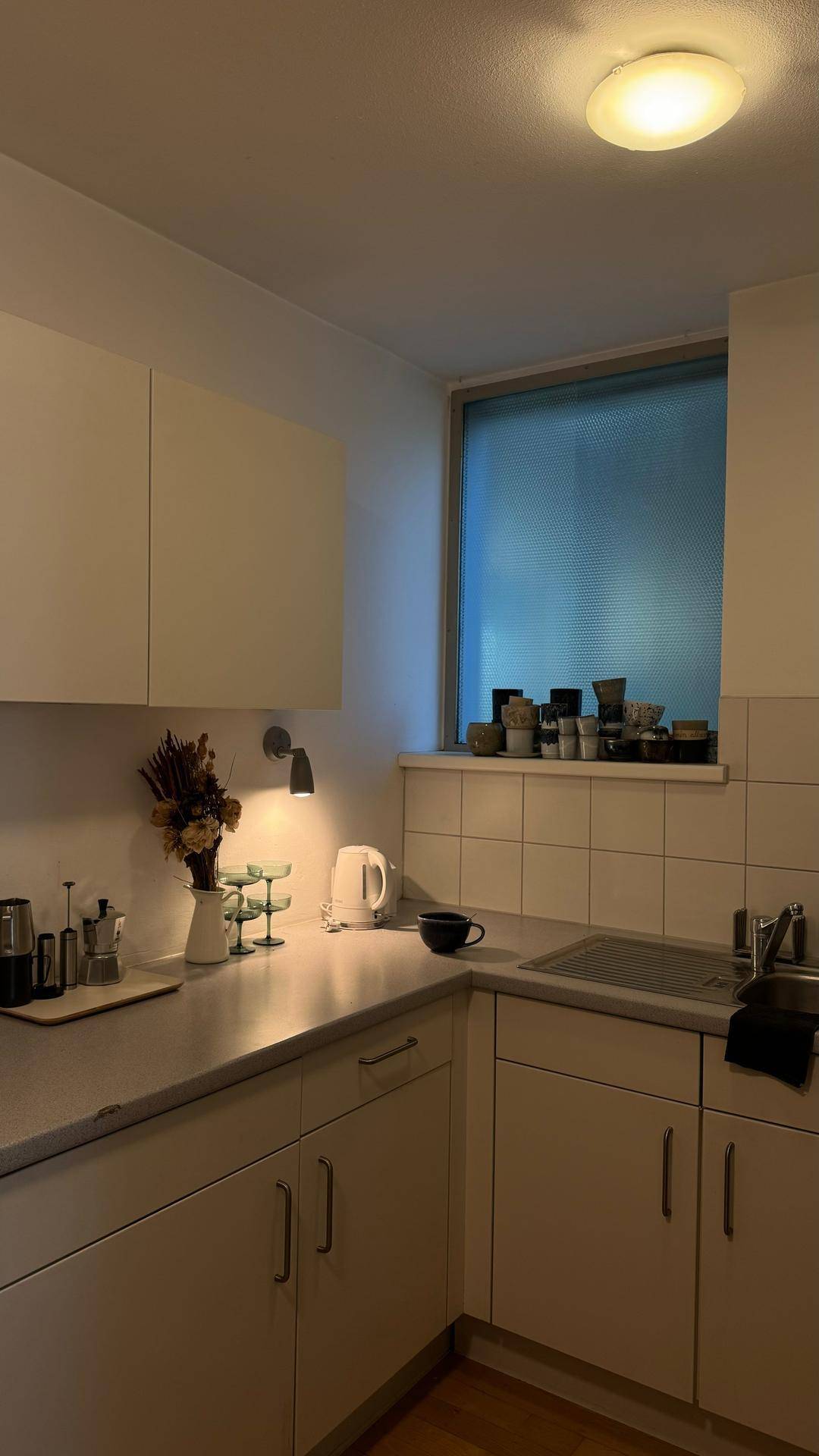 Funktionale Küche mit weißen Schränken, Spüle und einem Fenster mit Milchglas.
