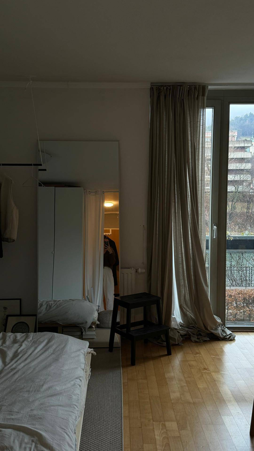Schlafzimmer mit Bett, großem Spiegel und Fenster mit Vorhängen und Stadtblick.