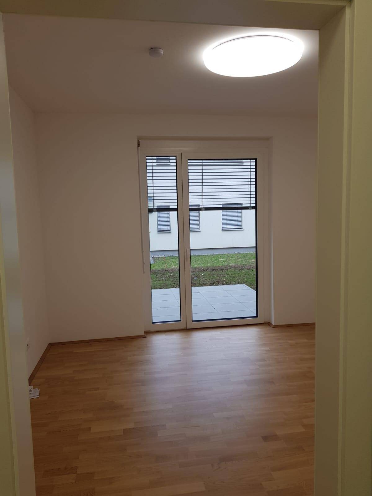 Leerer Raum mit Parkettboden und großer Fensterfront, die viel Tageslicht hereinlässt.