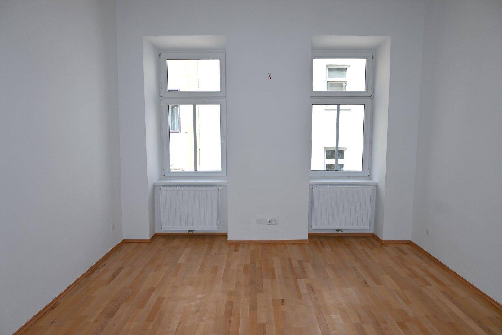 Geräumiges Zimmer mit zwei großen Fenstern, Parkettboden und weißen Wänden, sehr hell.