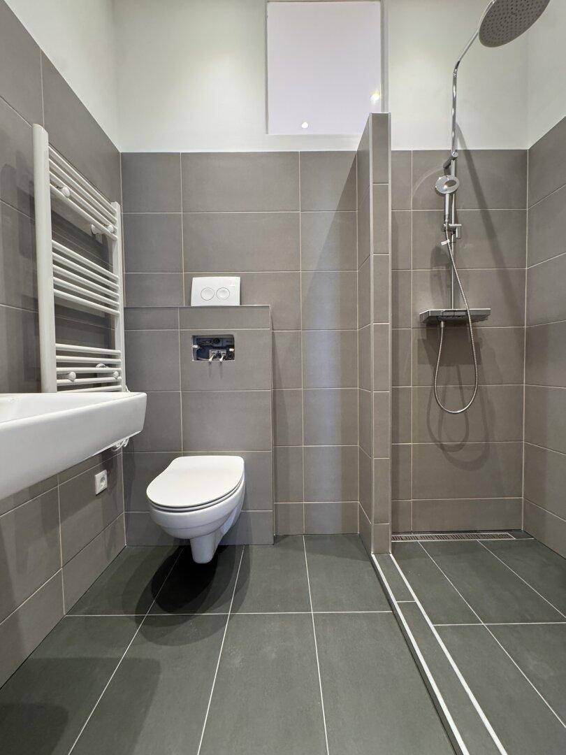 Modernes Badezimmer mit grauen Fliesen, ebenerdiger Dusche, WC und Waschbecken.