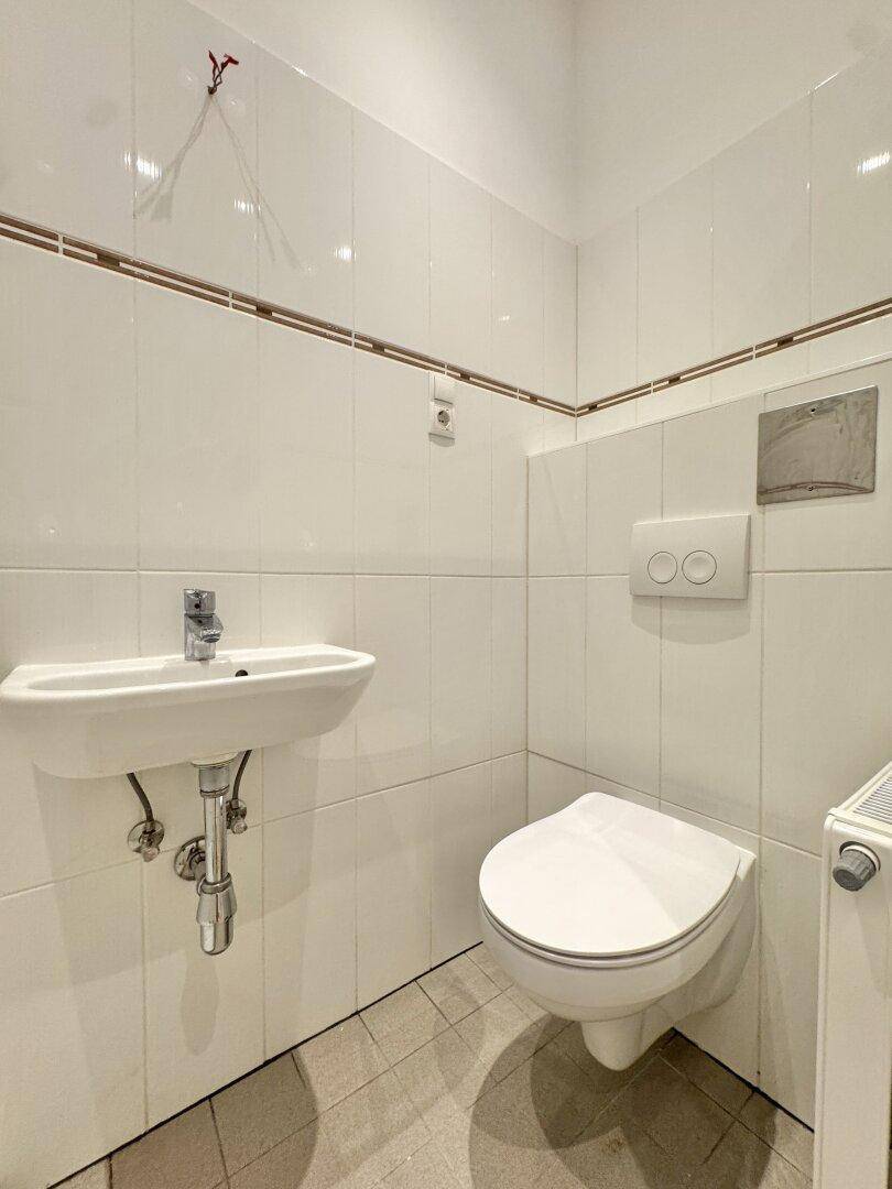 Kleines WC mit weißen Fliesen, wandhängender Toilette und einem Handwaschbecken.