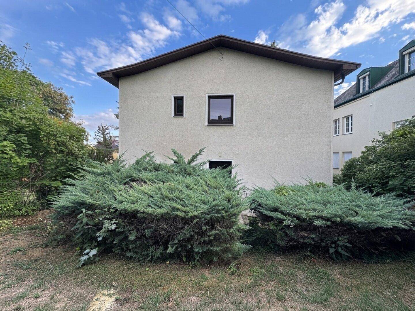 Rückseite des Hauses mit Gartenbereich und Sträuchern unter einem blauen Himmel.