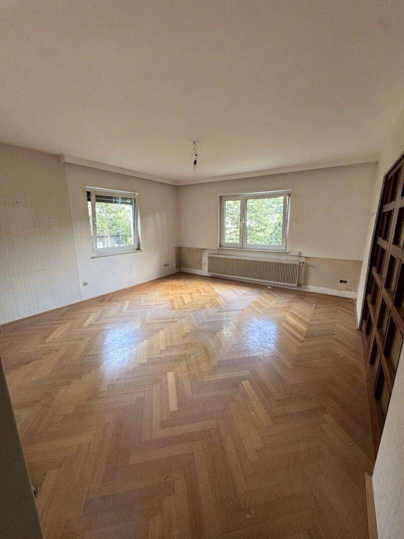 Geräumiges Wohnzimmer mit Fischgrätparkettboden und zwei großen Fenstern, bereit zur Renovierung.