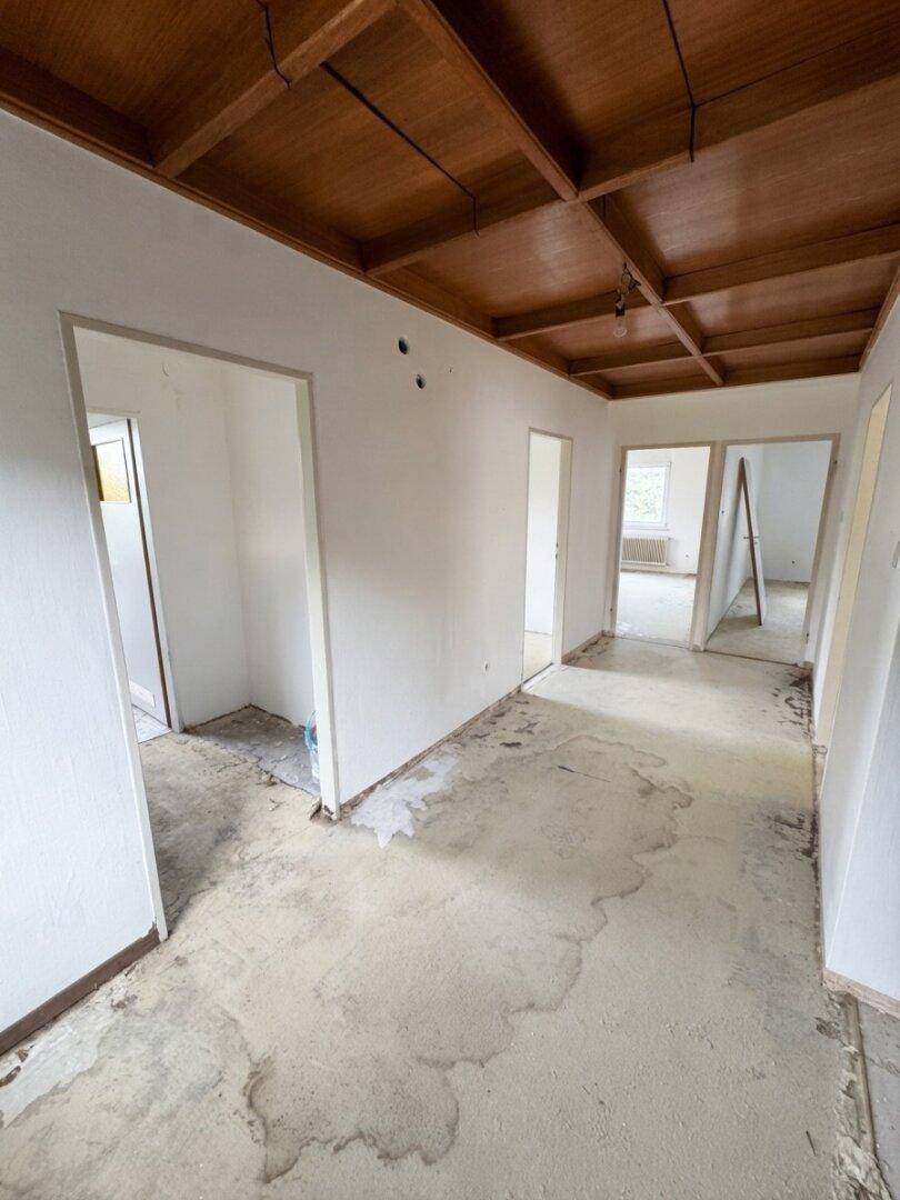Langer Flur mit Holzbalkendecke und teilweise entferntem Bodenbelag, führt zu mehreren Räumen.
