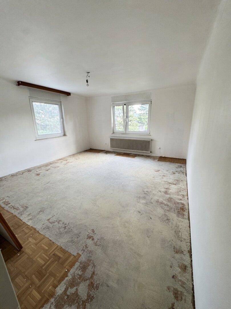 Helles Schlafzimmer mit teilweise entferntem Bodenbelag und zwei Fenstern, bereit zur Sanierung.