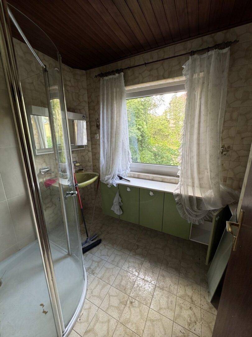 Badezimmer mit Duschkabine, Fenster und grünem Waschtischunterschrank, geflieste Wände.